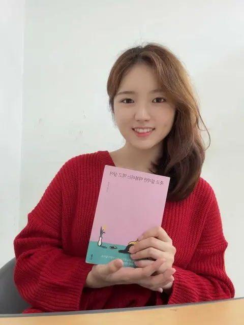 Jo Eun Bit in a red jumper smiles at the camera as she holds a copy of the book 'I Want to Die but I Want to Eat Tteokbokki' by Baek Se-hee.