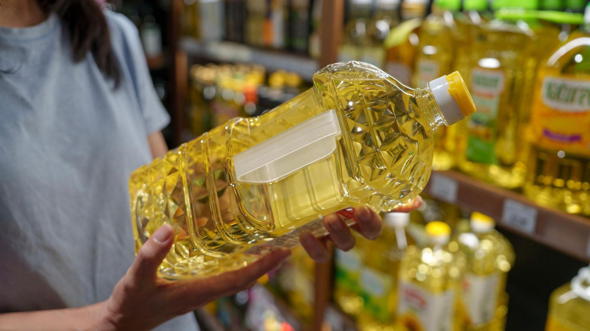 Close-up of a shopper holding a bottle of cooking oil in a supermarket aisle, focusing on food choices, daily shopping, and consumer lifestyle.