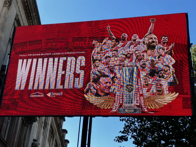 A large screen with a red background and a cut out of rugby players and a large silver trophy. The word winners is in capital letters/