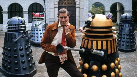 BBC Terry Nation wearing a red tie and tan jacket posing with four multi-coloured Daleks in front of a grand looking building