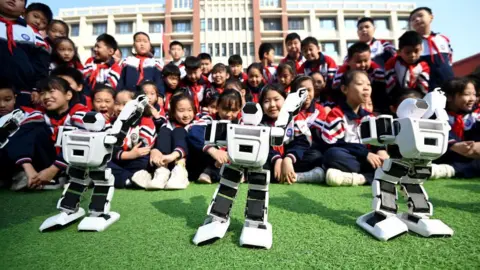 Getty Images Dozens of Chinese primary school students, dressed in school uniforms, sit on a playground watching three robots dance on the ground