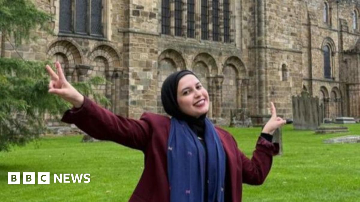 Sana el-Azab is sitting on a wall circling Durham Cathedral. She is smiling and is doing a peace sign with her hands.