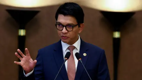 Reuters A man wearing black-rimmed glasses and in a blue suit with a red tie speaking into two microphones, holding up his hand as he speaks