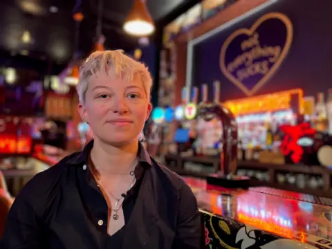 A woman with short blonde hair and a black shirt sits at a bar