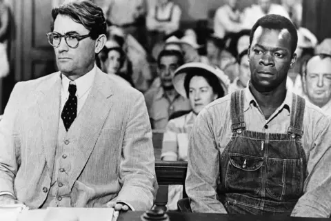 Getty Images Courtroom scene from the film of To Kill A Mockingbird shows Gregory Peck on the left seated in pale suit and waistcoat with glasses, and papers and hat on desk in front. On his right is Brock Peters who played Tom Robinson in dark dungarees and shirt. Both have serious expression on their faces.
Behind them people in the courtroom.