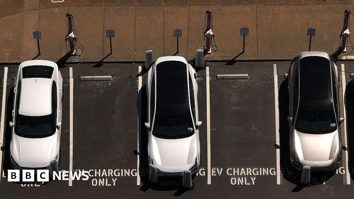 In an aerial view, three white electric cars sit parked at a charging station, with 'EV Charging Only' signs painted on the ground