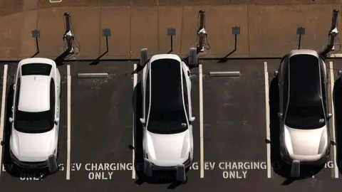 Getty Images In an aerial view, three white electric cars sit parked at a charging station, with 'EV Charging Only' signs painted on the ground