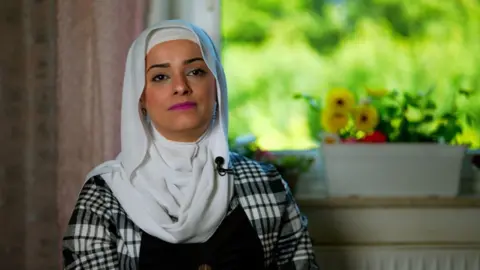 Headshot of a woman wearing a black and white checked top with a white headscarf. She is wearing pink lipstick. There is a pot with yellow flowers in the background.