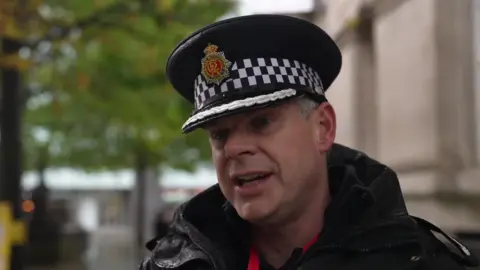 BBC Assistant Chief Constable Chris Sykes, wearing his black Greater Manchester Police cap and a large black jacket