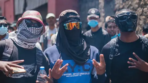 Gamma-Rapho via Getty Images Masked protesters wearing wearing black glasses and and caps pose on a street 