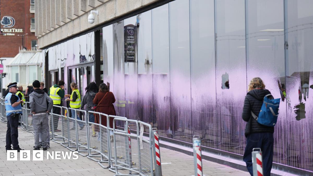A long window at the front of a building is spray-painted pink. Some panes have been smashed.