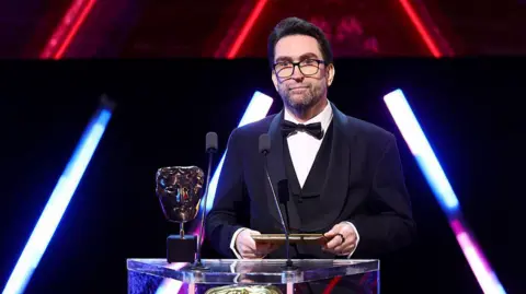 Getty Images A man wearing glasses and a three-piece suit with bow tie stands on-stage behind a lectern, holding an envelope in both hands. On the lectern are two microphones and a Bafta award - a golden trophy shaped like a theatrical mask.