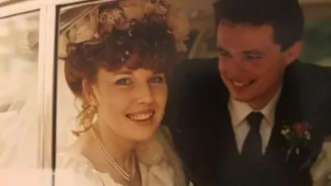 Liv Heeney A photo of a bride and groom in a car on their wedding day. The woman has wavy red hair tied up and is wearing a pearl necklace, floral headpiece and a white wedding dress and is smiling into the camera. The man has short brown hair and is wearing a dark suit and tie with a white shirt and floral pin. He is looking wistfully at the bride.