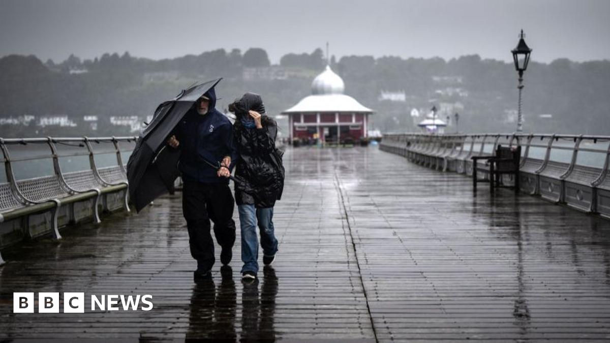 A couple walk on a pier while trying to shield themselves from the rain using an umbrella and their hooded coats.