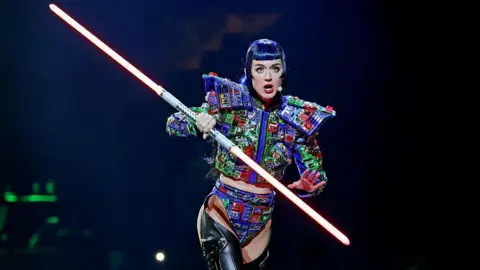 Getty Images Katy Perry is on stage and has a blue-black wig with a full fringe. She is wearing thigh-high black booths, a cropped jacket that is blue, green and red with large shoulder pads, and matching shorts.