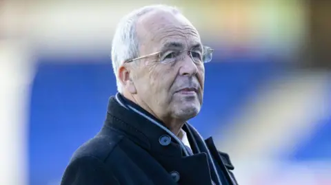 SNS Roy MacGregor is an older man with white hair and glasses. He is wearing a black, woollen coat and scarf and standing in the outdoors.