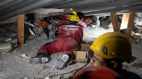 Getty Images Three people wearing yellow hard hats and red uniforms lay facing down under collapsed debris. The words 'FIRE RESCUE' are printed in white on one person's back. In the foreground is the head of another person wearing a yellow hard hat 