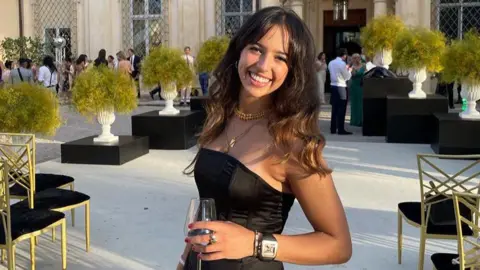 Supplied Grace O'Malley-Kumar smiling for the camera with long brown hair while holding a glass of wine at an outdoors event.