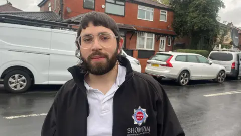 David Elkouby, who is an Orthodox Jew, stands in the middle of a road on a wet day wearing a black jacket and white polo shirt. 