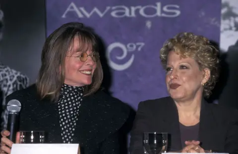 Ron Galella Collection via Getty Images Diane Keaton looking and smiling at Bette Midler, both sitting at a table