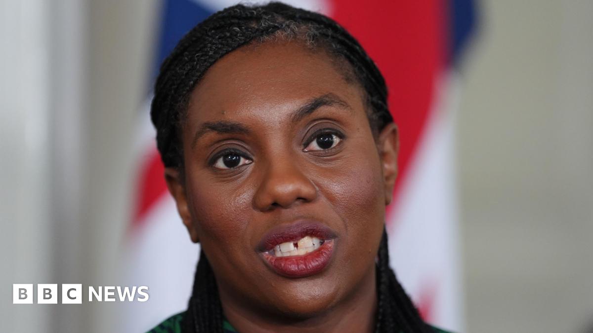 Close-up of Conservative Party leader Kemi Badenoch wearing a green patterned top. The blurred background features part of a Union Jack flag.