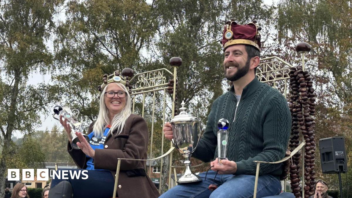 A woman and a man sitting on chairs that say Queen Conker and King Conker across the back. They are both holding silver trophies, wearing crowns and smiling. She has long grey hair and is wearing glasses, a brown coat and blue jeans. He has a dark beard and is wearing a green jumper and blue jeans.