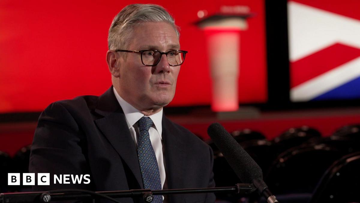 Sir Keir Starmer, dressed in a dark suit, white shirt, and blue patterned tie, is seated and speaking into a microphone during an interview at the Labour Party conference. Behind him is a red backdrop with blurred elements of the Union Jack and an indistinct podium. The auditorium setting includes rows of empty black chairs