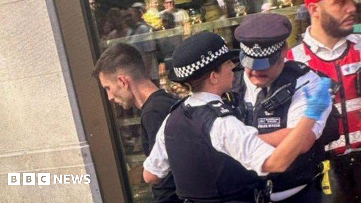 A white man with short dark hair and a black top and trousers stands with his hands behind his back as he is arrested by two officer, with a security guard in a red uniform in the background
