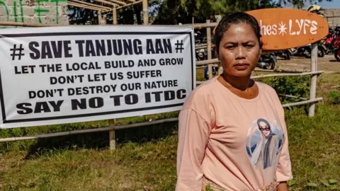 Just Finance International Ella Nurlaila in a peach coloured long-sleeved shirt, looking into the camera with a burrowed eyebrows. Behind her is a large banner that reads: "Save Tanjung Aan, let the local build the grow, don't let us suffer, don't destroy our nature, say no to ITDC".