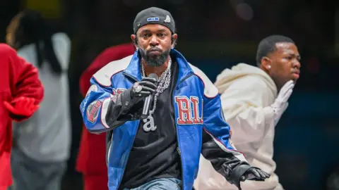 Getty Images Rapper Kendrick Lamar wears a blue varsity jacket and backwards baseball cap while performing in front of dancers at the 2025 Super Bowl half-time show.
