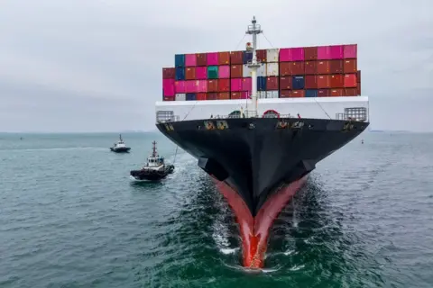 Getty Images A large cargo ship is sailing away from the camera. It is stocked with storage containers of bright pinks, reds, greens and blues.