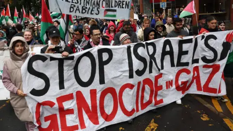 PA A large crowd, many holding Palestinian flags, marching along a street. At the front several people hold a large white banner with the words Stop Israel's Genocide in Gaza on it. 