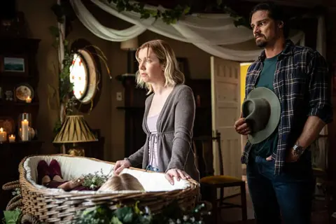 BBC/The Ridge Production Ltd The image depicts a solemn scene involving a woman and a man in a dimly lit room with a wicker casket. A wicker casket is central in the image, adorned with flowers and containing what appears to be a body covered with a blanket. The woman, Lyle, is leaning on the casket, possibly in a moment of reflection or farewell. The man stands nearby, holding a hat and looking toward the first individual.