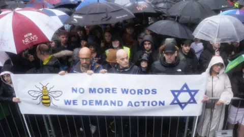 PA Media A large group of people standing behind railings and a banner that says 'No more words, we deman action', with a picture of a bee and a Star of David. People look serious and many are carrying umbrellas as it is raining heavily.