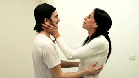 Israel Defense Forces Released Israeli hostage, Matan Zanguaker, holds his mother's waist, while she holds his neck and face with her hands. They are smiling at each other and are both wearing cream-coloured clothes.