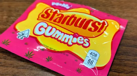 BBC A bright pick and yellow plastic packet with "Mini Starburst Gummies" written on it. There are small images of cannabis leaves in the background.