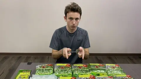 ViralPress Man in grey t-shit sat in front of a table. He is pointing at 10 packages in green foil. He is wearing handcuffs. He has short brown hair. 