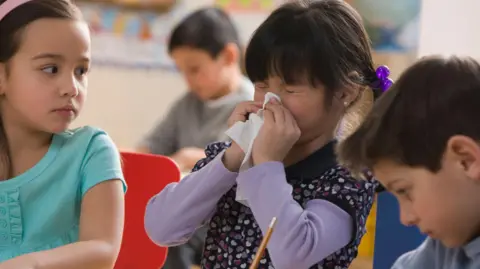 Getty Images A little girl sneezing into a tissue at nursery, while another child looks on