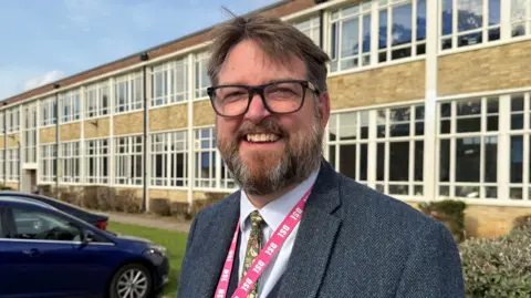 Jack Hunter/BBC Duncan Spalding, the Executive Headteacher of Aylsham High School who is standing outside the secondary school. He is looking directly at the camera and is smiling. He is wearing a white shirt with a colourful tie and a grey suit jacket over the top. He is also wearing a bright pink lanyard round his neck. He has brown hair and facial hair and is wearing a pair of black framed glasses.