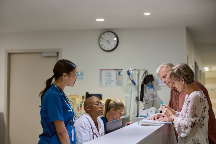 Senior couple filling form at hospital reception desk, medical staff offer support.