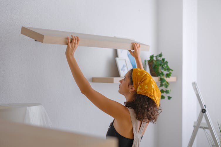 A woman in overalls installs a floating shelf on a white wall