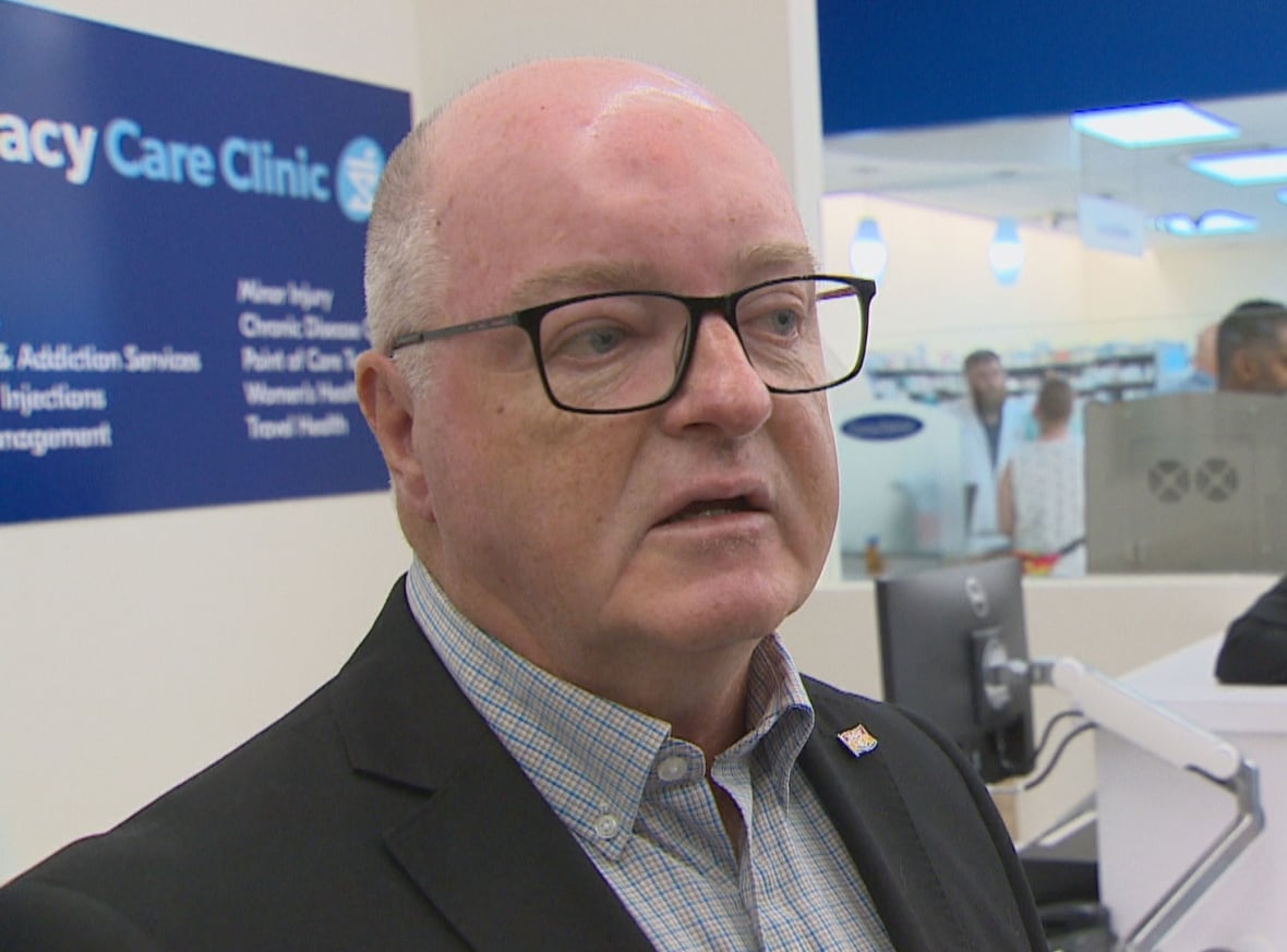 A man standing in a pharmacy, speaking, as others look on.
