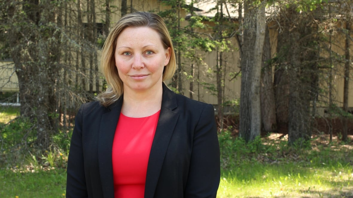 A woman in a red dress and black blazer poses for a picture in a park on a sunny day.