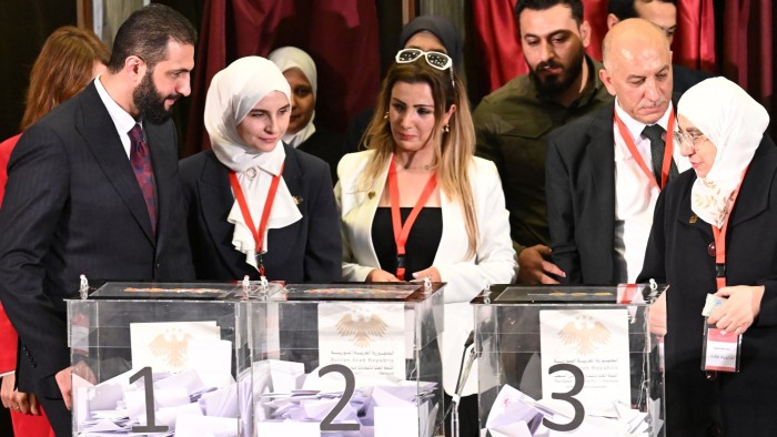 Ahmed al-Sharaa (left) observes as officials stand by ballot boxes labeled 1, 2, and 3.