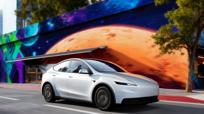 A white Tesla Model Y Standard driving past a colourful mural featuring a large orange planet and abstract space-themed designs.
