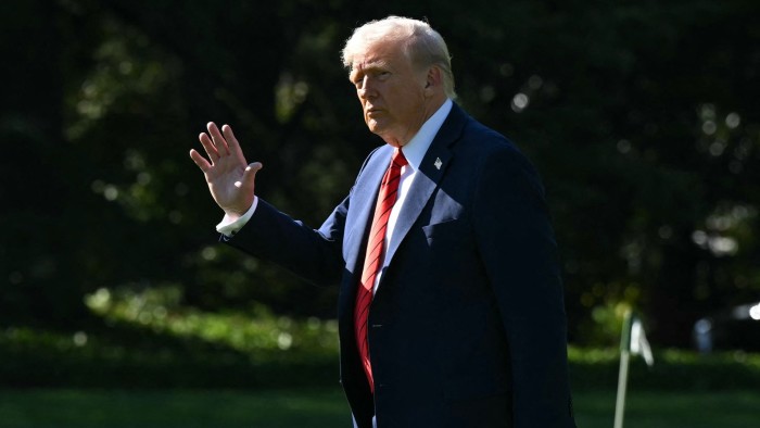 Donald Trump waves while walking outdoors in a suit and red striped tie.