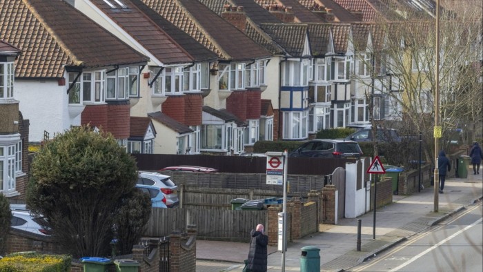 Rows of semi-detached houses with tiled roofs line a residential street in Greenwich, London. A few people walk along the sidewalk.