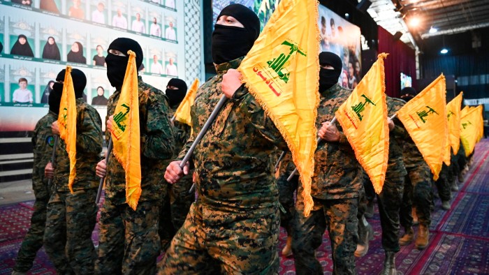 Masked men in camouflage uniforms march indoors holding yellow Hezbollah flags, with portraits of people displayed on the wall behind them.
