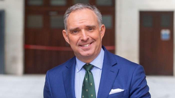 Mark Sedwill smiles while standing outdoors in a blue suit and green tie.