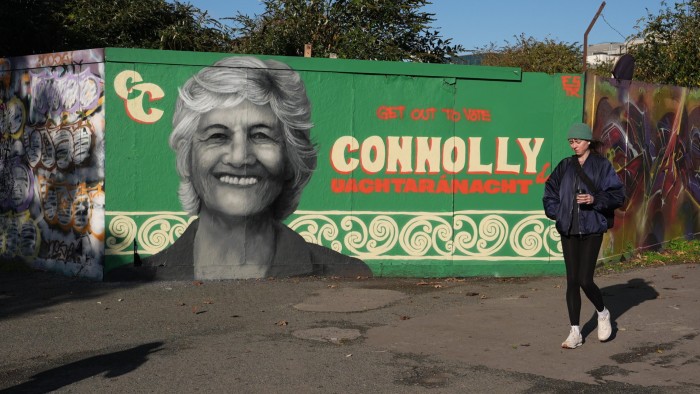 A mural of Catherine Connolly with a large smiling portrait and campaign text, as a woman walks by holding a coffee.